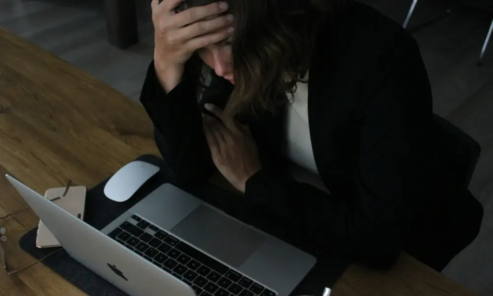Adult professional sitting at desk with head in hands, overwhelmed in front of laptop.