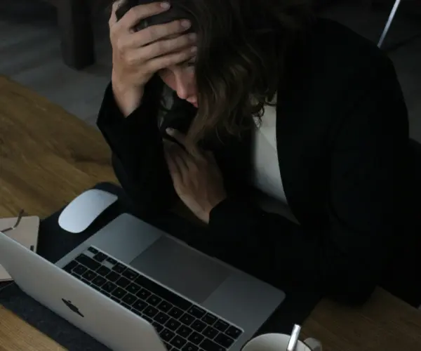 Adult professional sitting at desk with head in hands, overwhelmed in front of laptop.